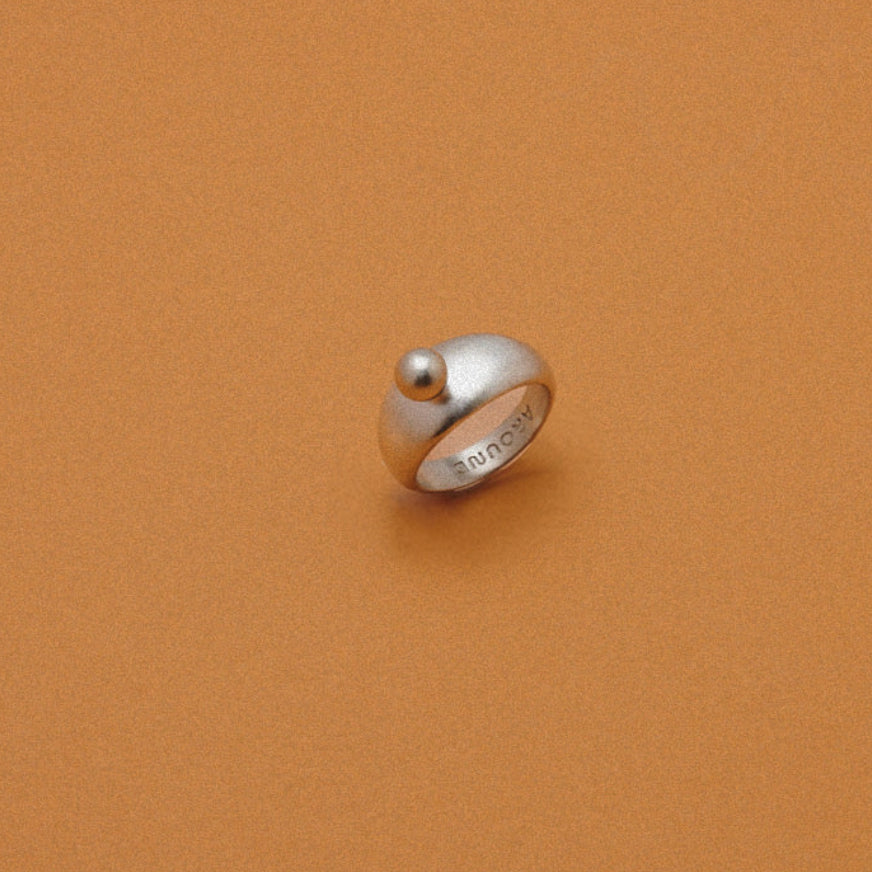 A silver ring with a small spherical bead on top, photographed on a solid warm brown background.