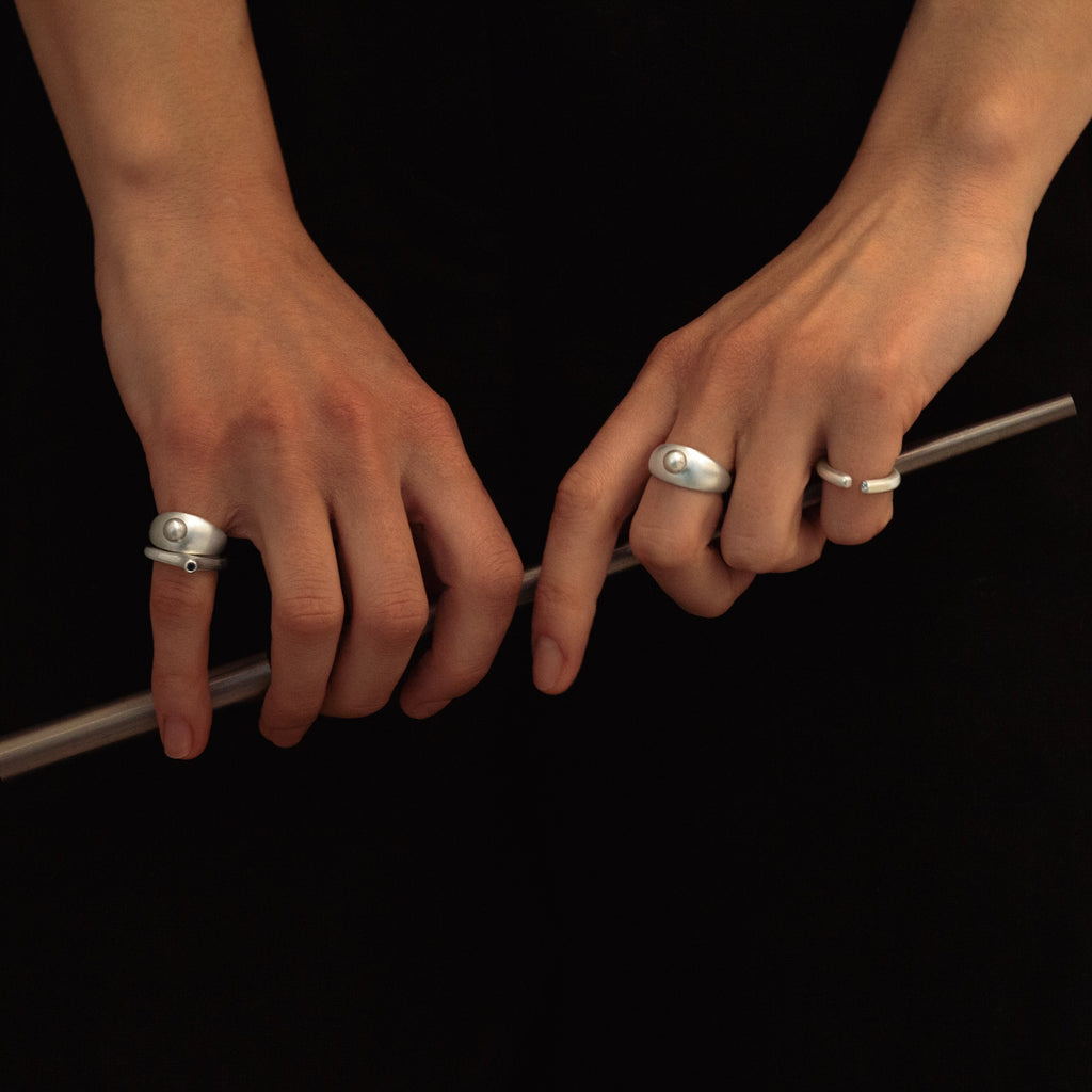 A close-up of a person's hands wearing several silver rings, including a large oval ring on the index finger and a thin band on the middle finger, while holding a metallic rod against a black dress.