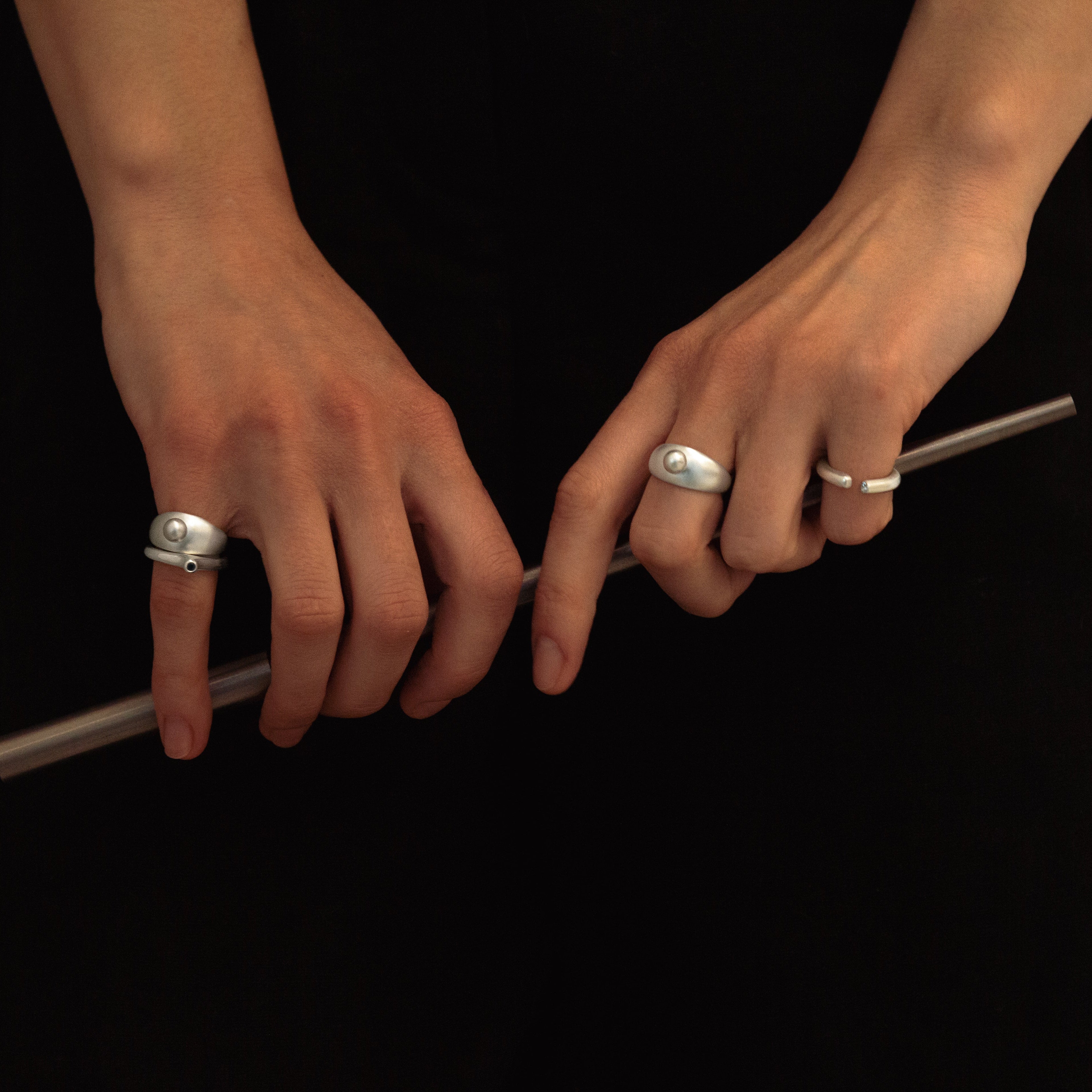 A close-up of a person's hands wearing several silver rings, including a large oval ring on the index finger and a thin band on the middle finger, while holding a metallic rod against a black dress.